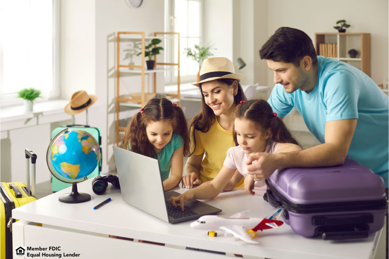 Photo of a family of four gathered around a desk looking at a computer screen. On the desk is a globe, airplane and luggage, giving the impression of trip planning.