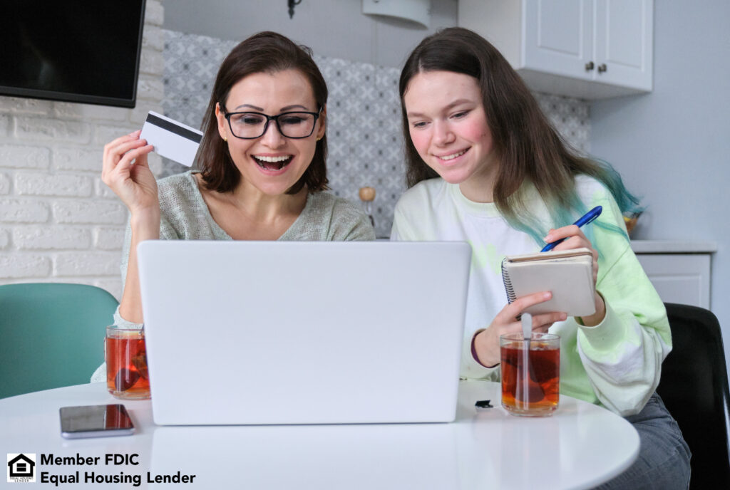 A mom and daughter sitting in from of a computer with the daughter taking notes and her mom holding a credit card. A financial responsibility teaching moment.