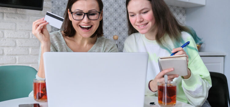 A mom and daughter sitting in from of a computer with the daughter taking notes and her mom holding a credit card. A financial responsibility teaching moment.
