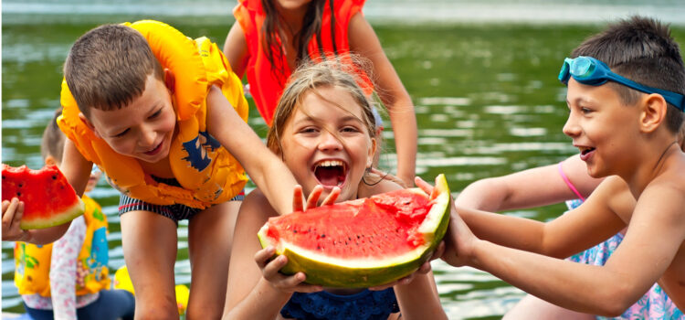 Here’s to the Ordinary Summer of 2026 Kids in lifejackets on a lake dock. Centermost child is holding a large slice of watermelon, a summer staple.